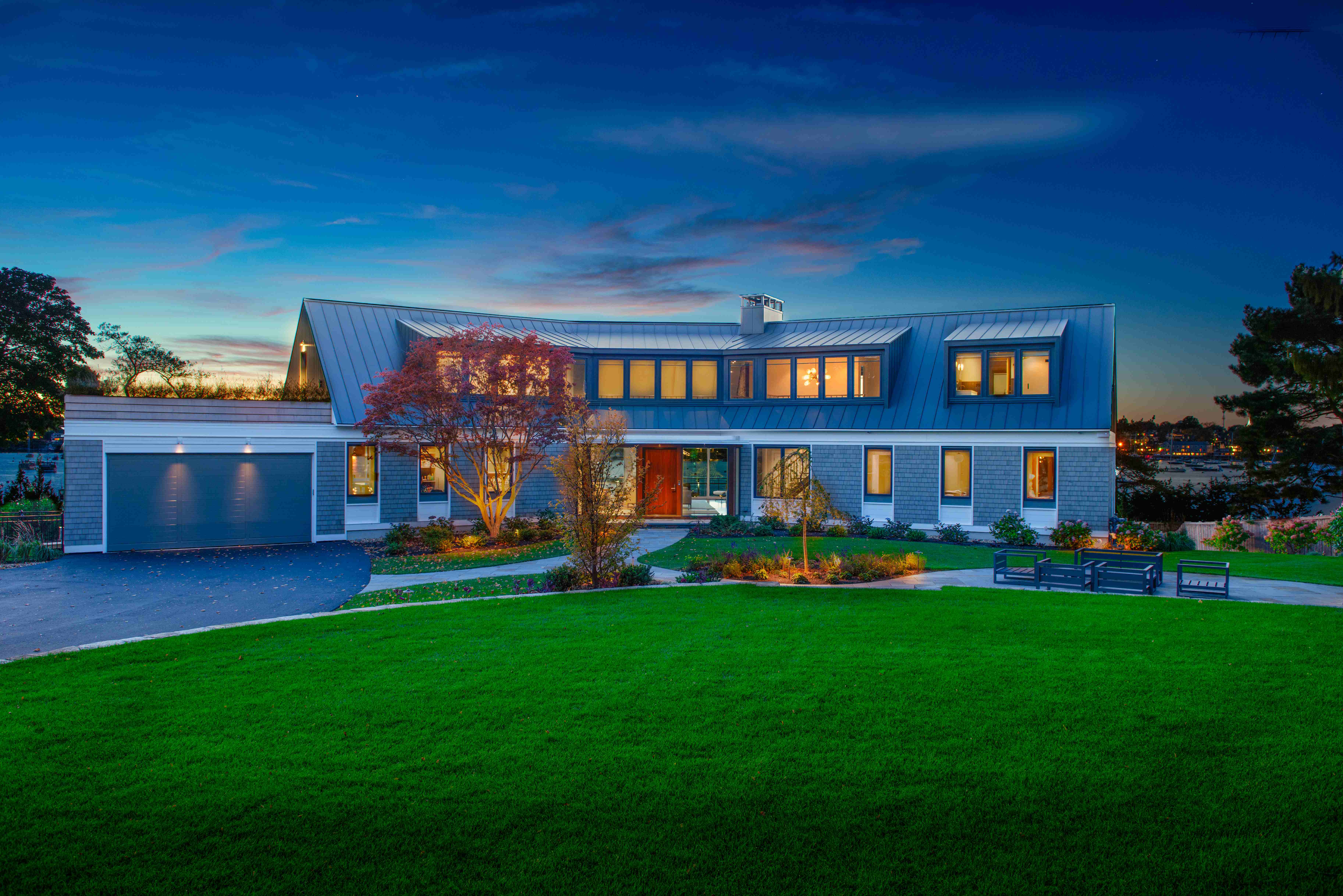 A newly constructed shingle-style coastal home in Marblehead, Massachusetts, glowing warmly at twilight beneath a standing-seam metal roof